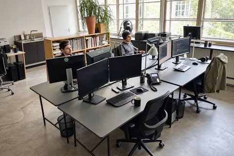 Zwei Kollegen arbeiten an Schreibtischen mit mehreren Monitoren in unserem hellen Büro.